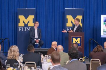 A master of ceremonies gestures while addressing a guest lecturer seated on stage to the left. Two banners displaying the University of Michigan-Flint logo are in the background.