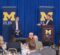 A master of ceremonies gestures while addressing a guest lecturer seated on stage to the left. Two banners displaying the University of Michigan-Flint logo are in the background.