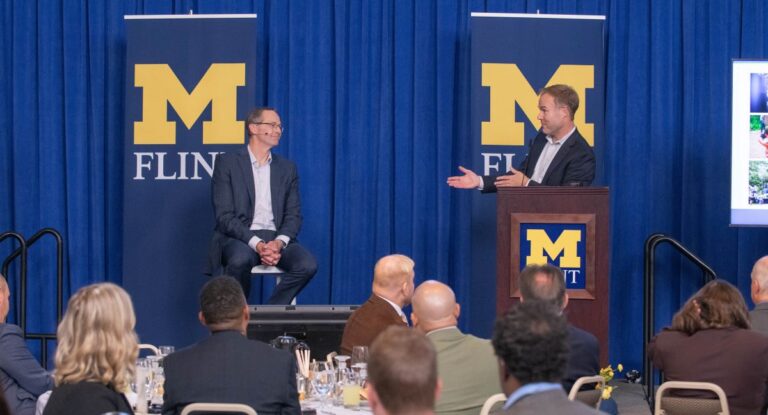 A master of ceremonies gestures while addressing a guest lecturer seated on stage to the left. Two banners displaying the University of Michigan-Flint logo are in the background.