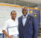 A young man in a white polo shirt holds a scholarship award certificate, standing next to a man in a blue suit and bow tie. They are smiling in a classroom setting with a blue and yellow "M" logo in the background.