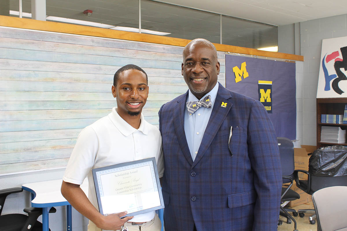 A young man in a white polo shirt holds a scholarship award certificate, standing next to a man in a blue suit and bow tie. They are smiling in a classroom setting with a blue and yellow "M" logo in the background.