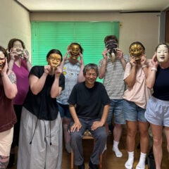 A group of eight people, some wearing traditional masks, pose together in a room. A man sits in the center, while others stand around him, holding masks to their faces.