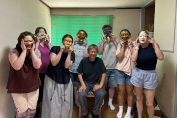 A group of eight people, some wearing traditional masks, pose together in a room. A man sits in the center, while others stand around him, holding masks to their faces.