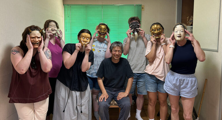 A group of eight people, some wearing traditional masks, pose together in a room. A man sits in the center, while others stand around him, holding masks to their faces.