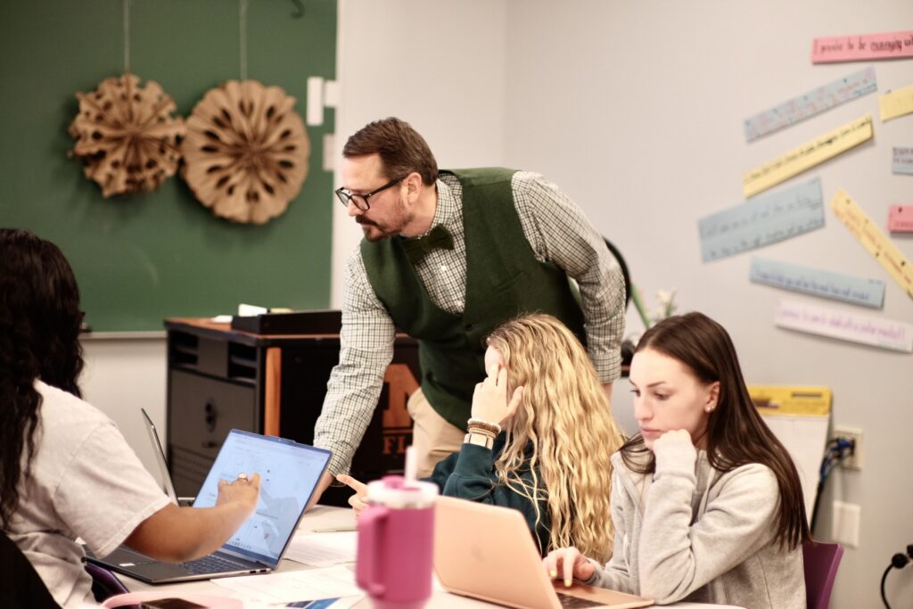A teacher in a green vest assists students at a table with laptops in a classroom, with educational materials on the walls.