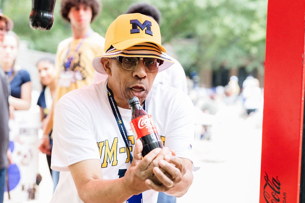 775307655_1508861147936482_3693205697734369597_n Chancellor Alexander wearing a yellow Michigan hat and glasses is holding a Coca-Cola bottle, looking focused. People are visible in the background.