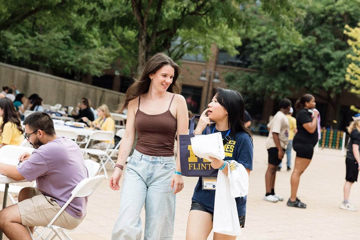 775333641_1508860617936535_852545463030378669_n Two young women walk together outdoors, one holding a folder that says "UM-FLINT." In the background, several people are seated at tables.