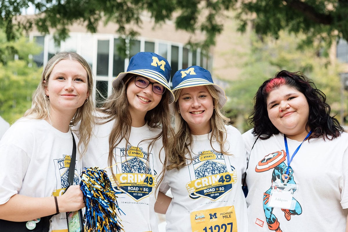 775375605_1508860944603169_2395469175870144014_n Four young students are smiling together outdoors, wearing matching white CRIM t-shirts and blue hats with a yellow "M." They are participating in an event, holding a pom-pom and wearing name tags.