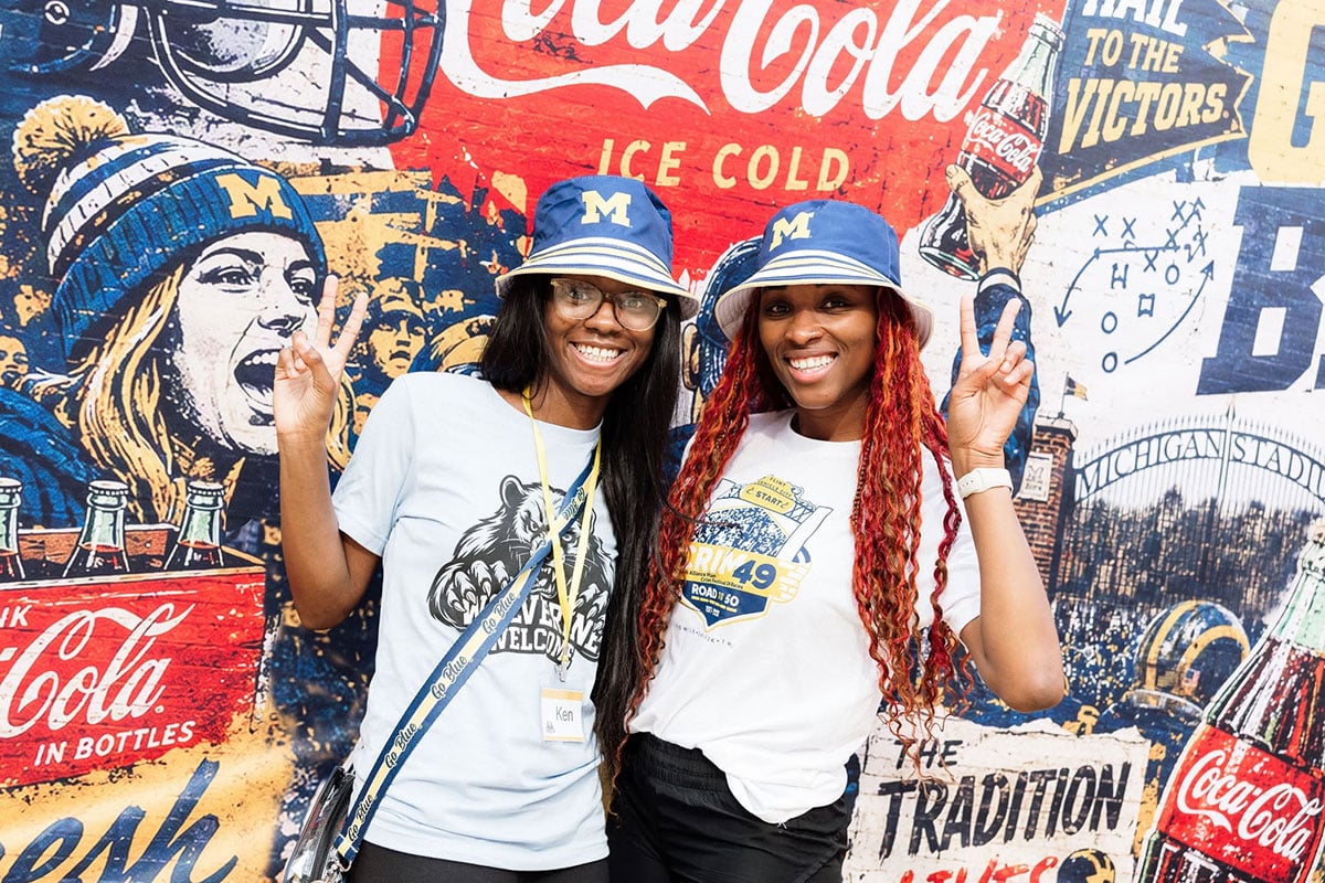 775375614_1508860584603205_7857019643411418234_n Two women pose for a photo, smiling and making peace signs, in front of a colorful mural featuring Coca-Cola branding and football imagery. One woman wears a blue and white bucket hat and a t-shirt with a bear graphic, while the other wears a white t-shirt and a blue bucket hat.
