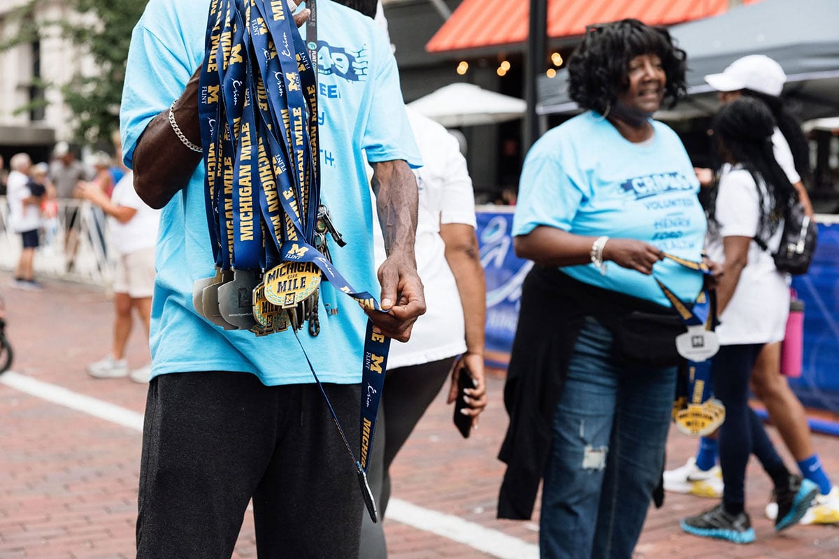 778869912_1508861144603149_7969107200036530584_n A person in a blue shirt holds multiple medals with "MICHIGAN MILE" written on them, while another person in a blue shirt stands nearby.