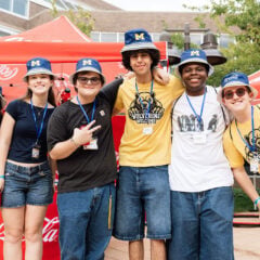 A group of six young adults poses together outdoors, wearing blue and yellow clothing and hats with an "M" logo. They are smiling and standing in front of a red Coca-Cola tent.