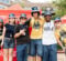 A group of six young adults poses together outdoors, wearing blue and yellow clothing and hats with an "M" logo. They are smiling and standing in front of a red Coca-Cola tent.