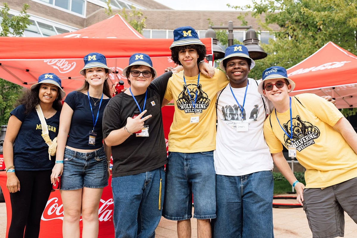 A group of six young adults poses together outdoors, wearing blue and yellow clothing and hats with an "M" logo. They are smiling and standing in front of a red Coca-Cola tent.