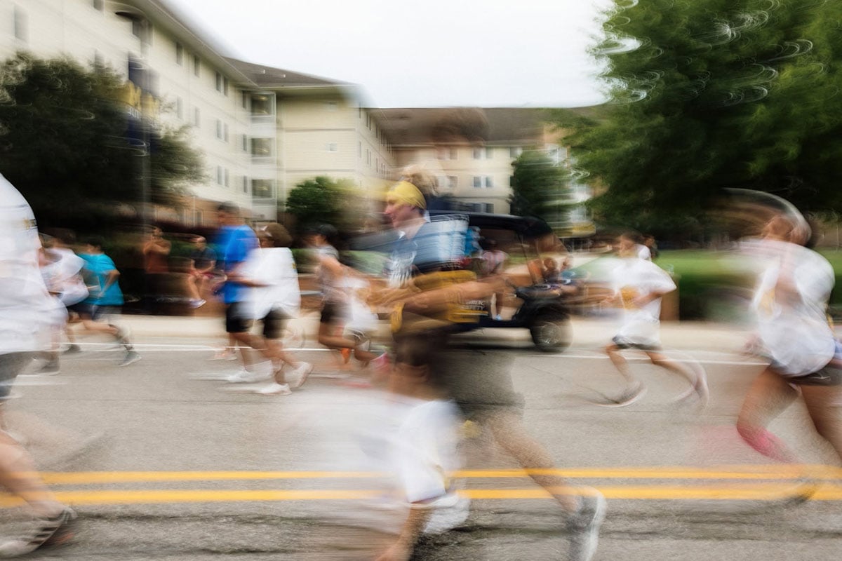 780157396_1508860637936533_2648267374185275386_n A blurred image of a group of people running on a street, with buildings and trees in the background.
