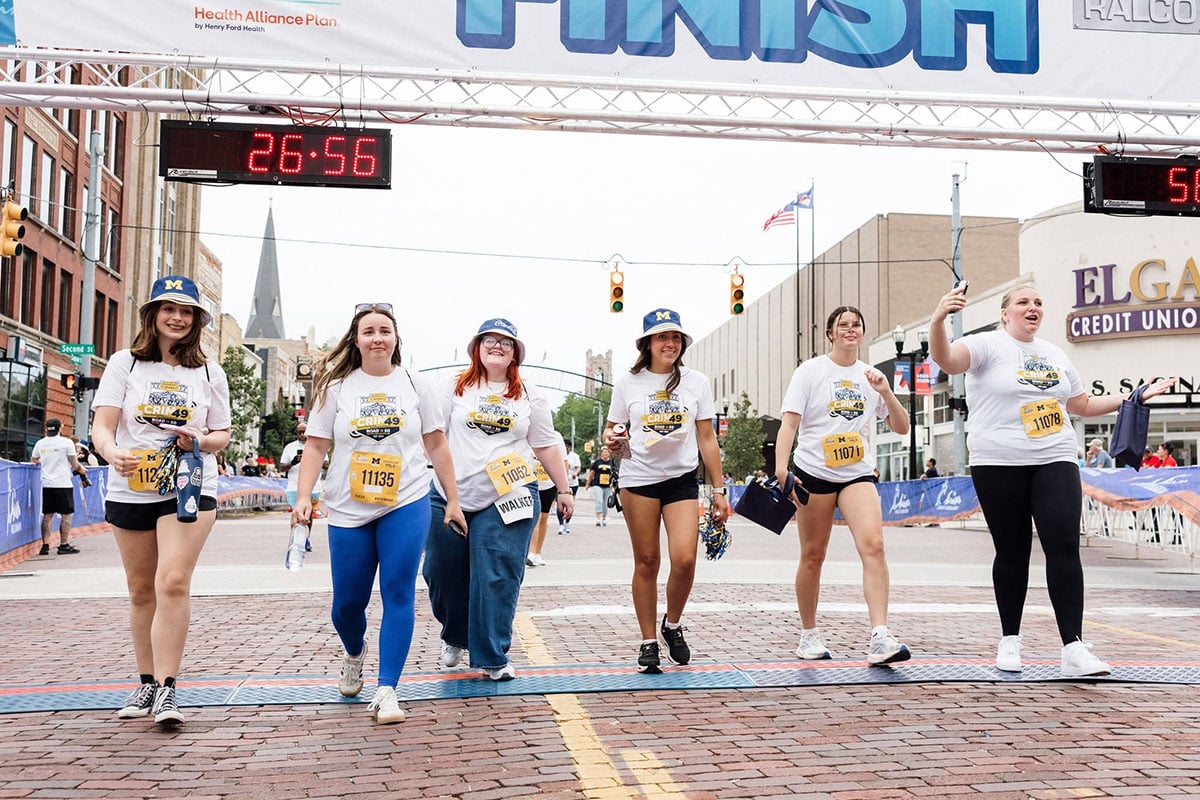 780672403_1508860951269835_2510586521377776754_n A group of six women in matching t-shirts and race bibs walk across a finish line, with a timer showing 26:56. They are smiling and celebrating.