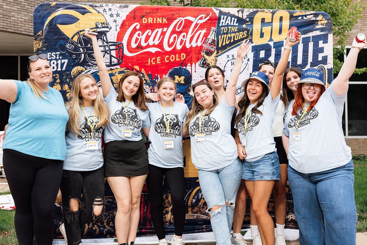 780909947_1508860634603200_4241032822845557473_n A group of ten young women poses together, smiling and celebrating in front of a colorful backdrop featuring Coca-Cola branding and a football theme. They are wearing matching light blue shirts that say "Wolverine Welcome."