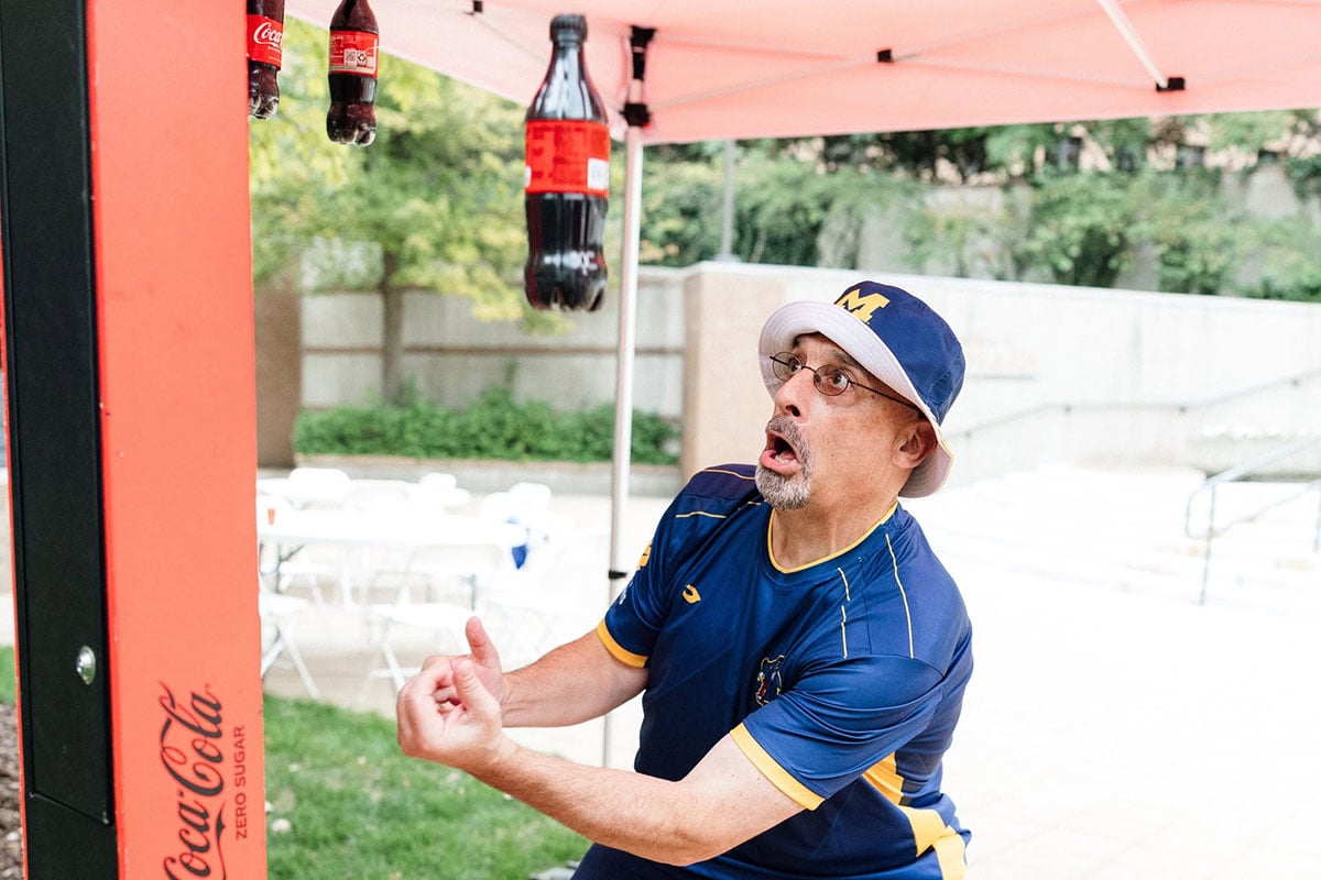 780981729_1508860864603177_4863098895487993669_n A man in a blue sports shirt and white bucket hat looks surprised while reaching for a bottle of Coca-Cola hanging above him.