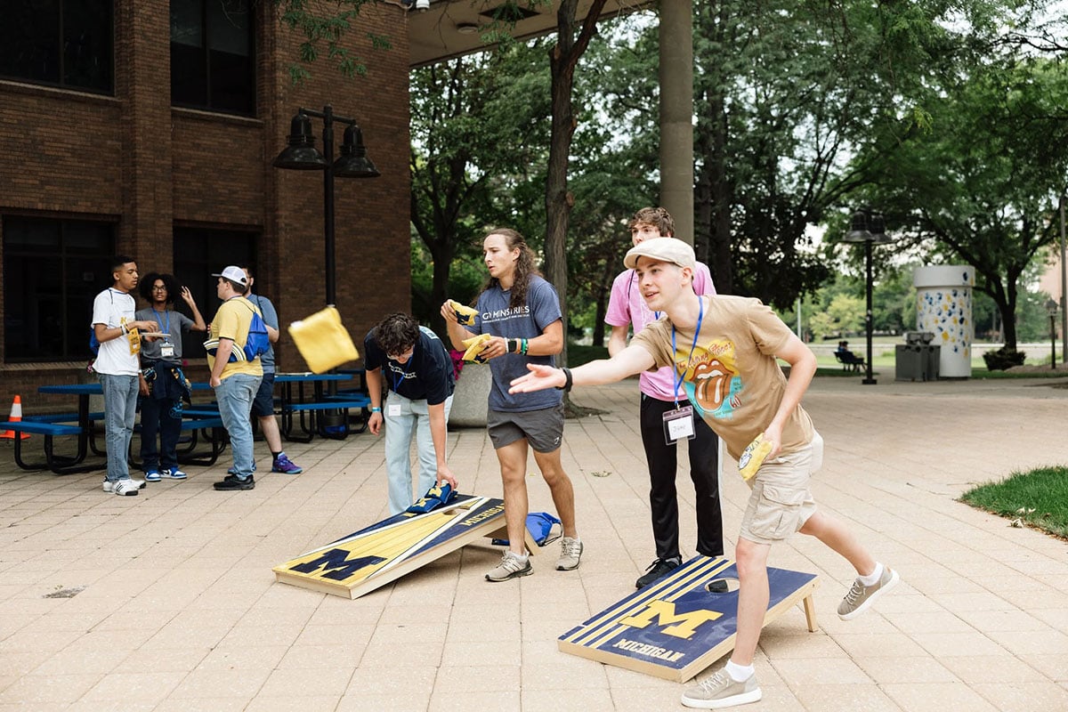 782145023_1508860881269842_1794393248914749575_n A group of young people are playing cornhole outside, with one person throwing a bean bag while others watch and chat.