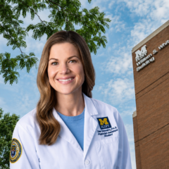A smiling woman in a white lab coat stands in front of a brick building with the sign "William S. White Building." She has long brown hair and is wearing a blue shirt underneath her lab coat.