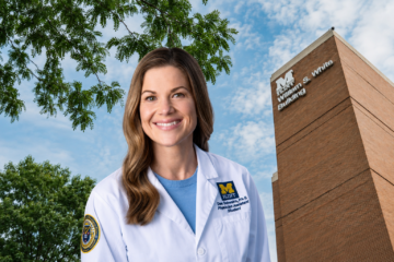 A smiling woman in a white lab coat stands in front of a brick building with the sign "William S. White Building." She has long brown hair and is wearing a blue shirt underneath her lab coat.