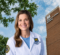 A smiling woman in a white lab coat stands in front of a brick building with the sign "William S. White Building." She has long brown hair and is wearing a blue shirt underneath her lab coat.