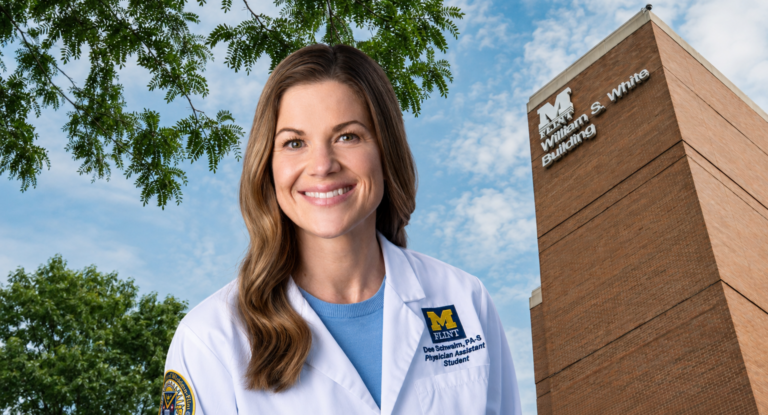 A smiling woman in a white lab coat stands in front of a brick building with the sign "William S. White Building." She has long brown hair and is wearing a blue shirt underneath her lab coat.