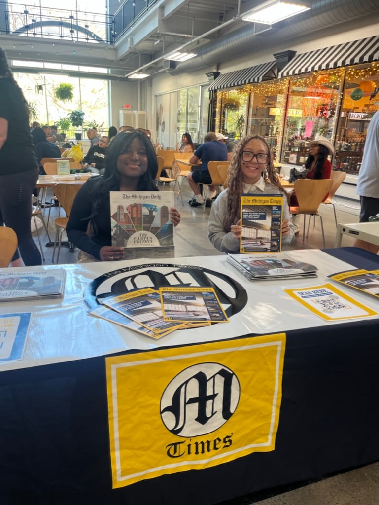 Two students are sitting at a table displaying various issues of "The Michigan Daily" newspaper. They are smiling and holding up copies of the newspaper. The table has a logo and additional printed materials.