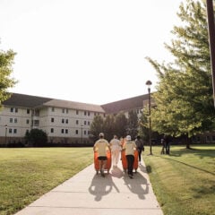 A group of people walking on a path in a grassy area, with a multi-story building in the background and trees lining the path.