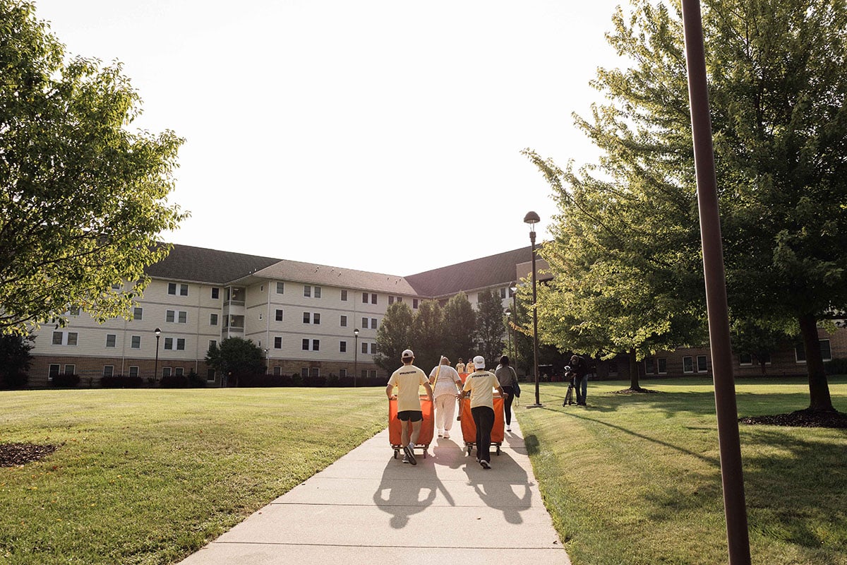 A group of people walking on a path in a grassy area, with a multi-story building in the background and trees lining the path.