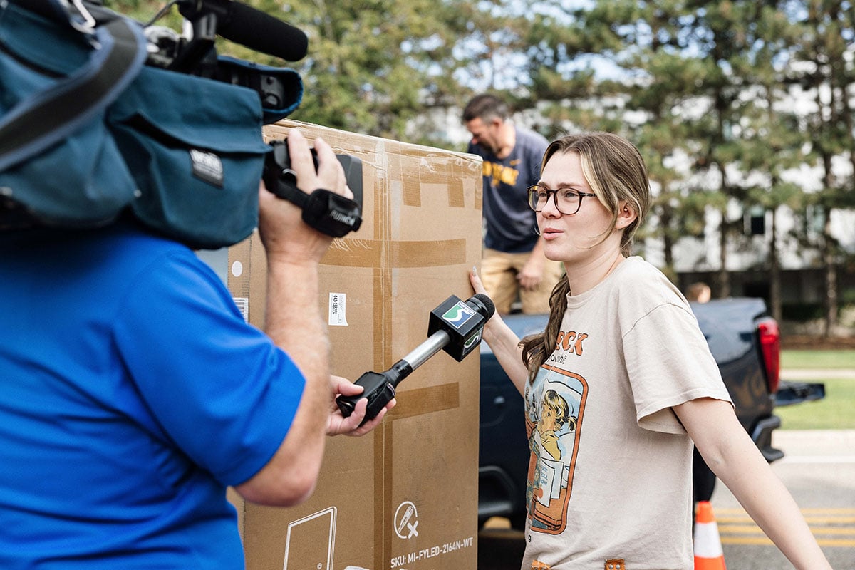 FLINT_2026 Move-In Day 0003 A woman with glasses speaks to a reporter holding a microphone, while a man is seen in the background unloading a large box from a truck.