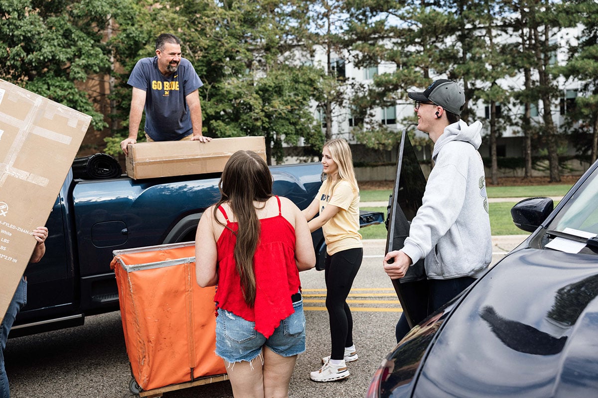 FLINT_2026 Move-In Day 0004 A group of four people are unloading a truck, carrying boxes and a cart. They appear to be helping with a move, smiling and interacting.
