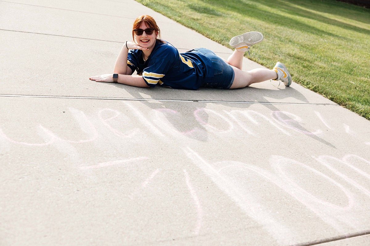 FLINT_2026 Move-In Day 0010 A person with red hair and sunglasses lies on a sidewalk, smiling next to chalk text that says "welcome home."