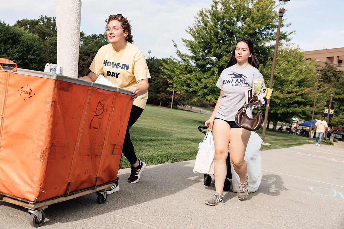 FLINT_2026 Move-In Day 0014 Two young women are moving items on a college campus. One pushes an orange cart labeled "MOVE-IN DAY," while the other carries bags and a drink.