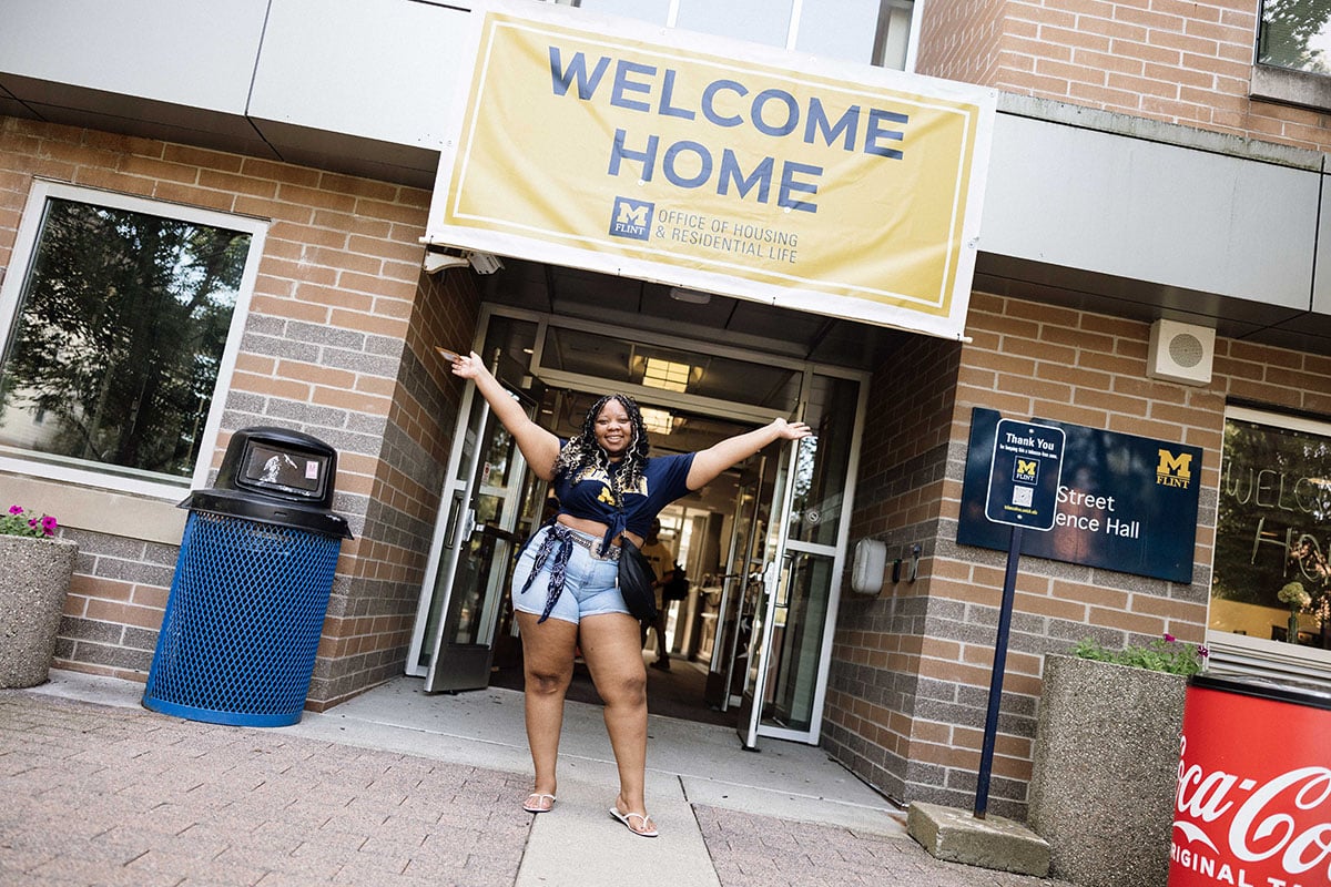 FLINT_2026 Move-In Day 0016 A person stands in front of a building entrance, smiling and gesturing with arms outstretched. A large banner above reads "WELCOME HOME."