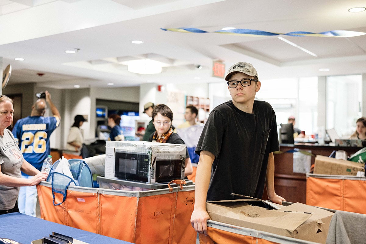 FLINT_2026 Move-In Day 0025 A young man in a black t-shirt and glasses stands next to a cart filled with boxes and a microwave, while people are seen in the background, some carrying items.