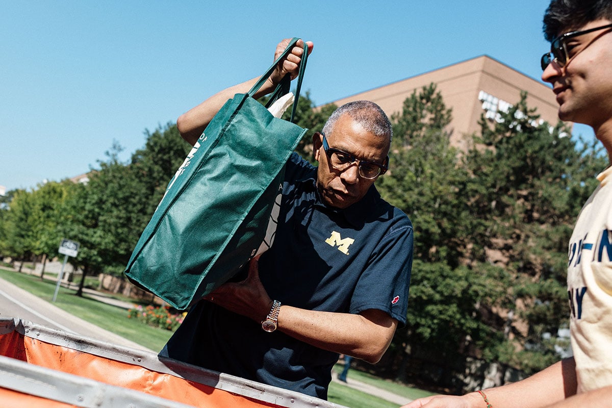 FLINT_2026 Move-In Day 0026 Chancellor Alexander wearing glasses and a navy shirt with a yellow "M" logo is lifting a green tote bag while a younger man watches. They are outdoors with trees and buildings in the background.