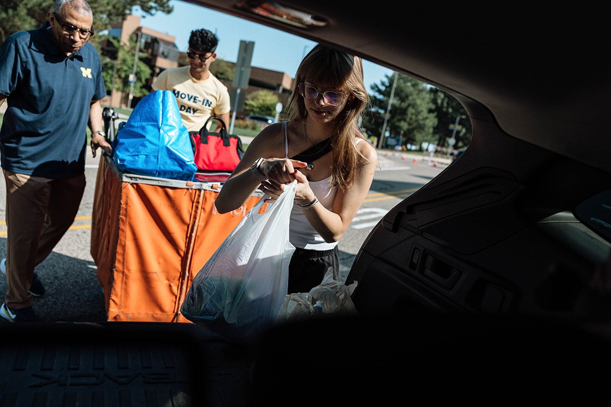 FLINT_2026 Move-In Day 0030 A young woman with long hair and glasses is unloading a plastic bag from a car, while a man in a blue shirt and another man in a tan shirt stand nearby with bags and a cart.