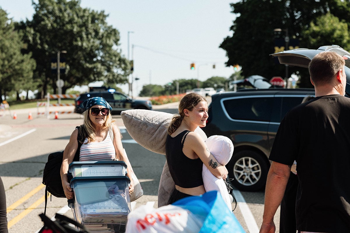 FLINT_2026 Move-In Day 0031 Two women are carrying items on a street, with one holding a large stuffed animal and the other pushing a cart filled with boxes. A car is parked nearby, and police vehicles are visible in the background.