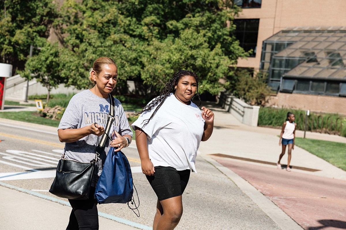 FLINT_2026 Move-In Day 0035 A woman and a girl walk together on a sunny day, with trees and a building in the background. Another girl is walking in the distance.