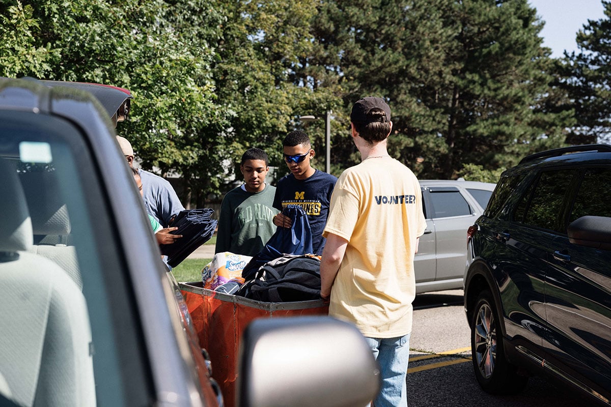 FLINT_2026 Move-In Day 0036 A group of young people is gathered around the back of a vehicle, unloading items. One person is wearing a yellow shirt with "VOLUNTEER" printed on it. Trees are visible in the background.