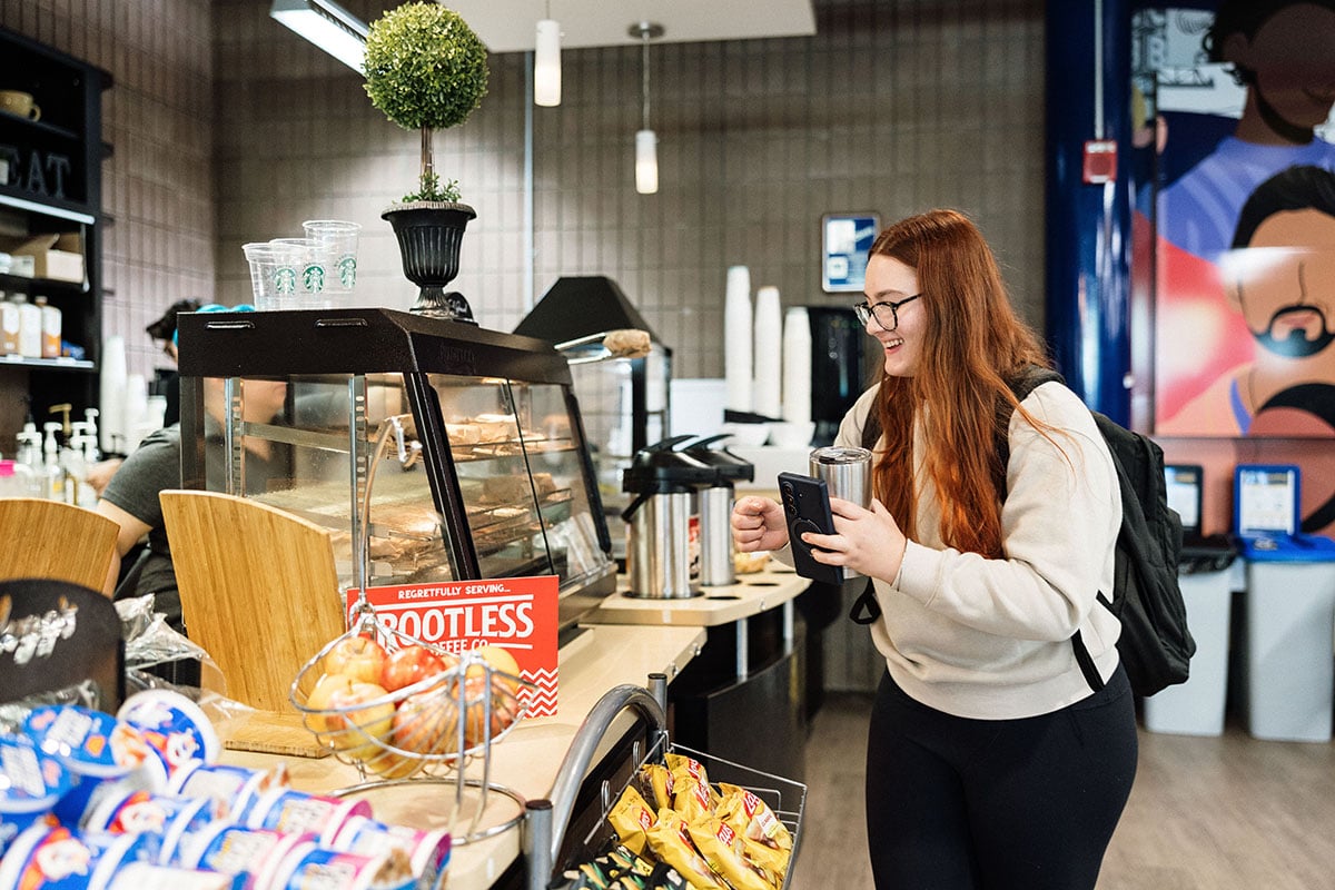 FLINT_First Day of Class Fall 2026_0002 A young woman with long red hair and glasses smiles while holding a phone and a travel mug in a coffee shop. She stands in front of a display case with baked goods and a sign that reads "Regrettably Serving: ROOTLESS Coffee Co." There are snacks and fruits in baskets nearby.