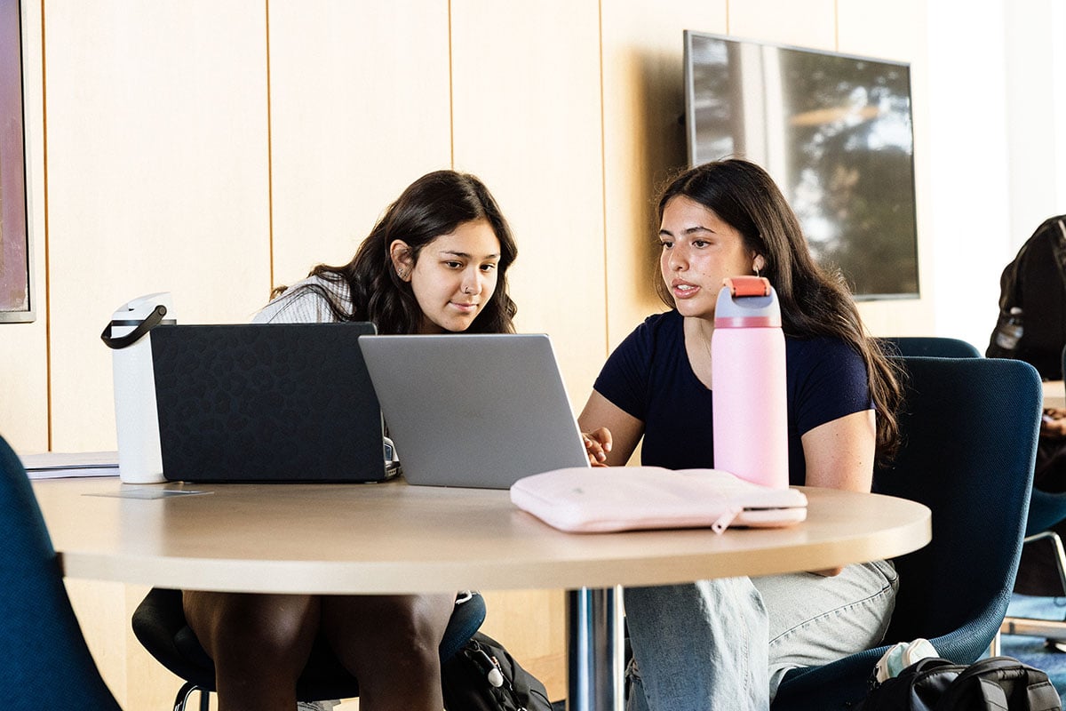 Two young women are sitting at a round table, focused on a laptop. One woman is pointing at the screen while the other looks on. A pink water bottle and a light pink laptop sleeve are on the table.