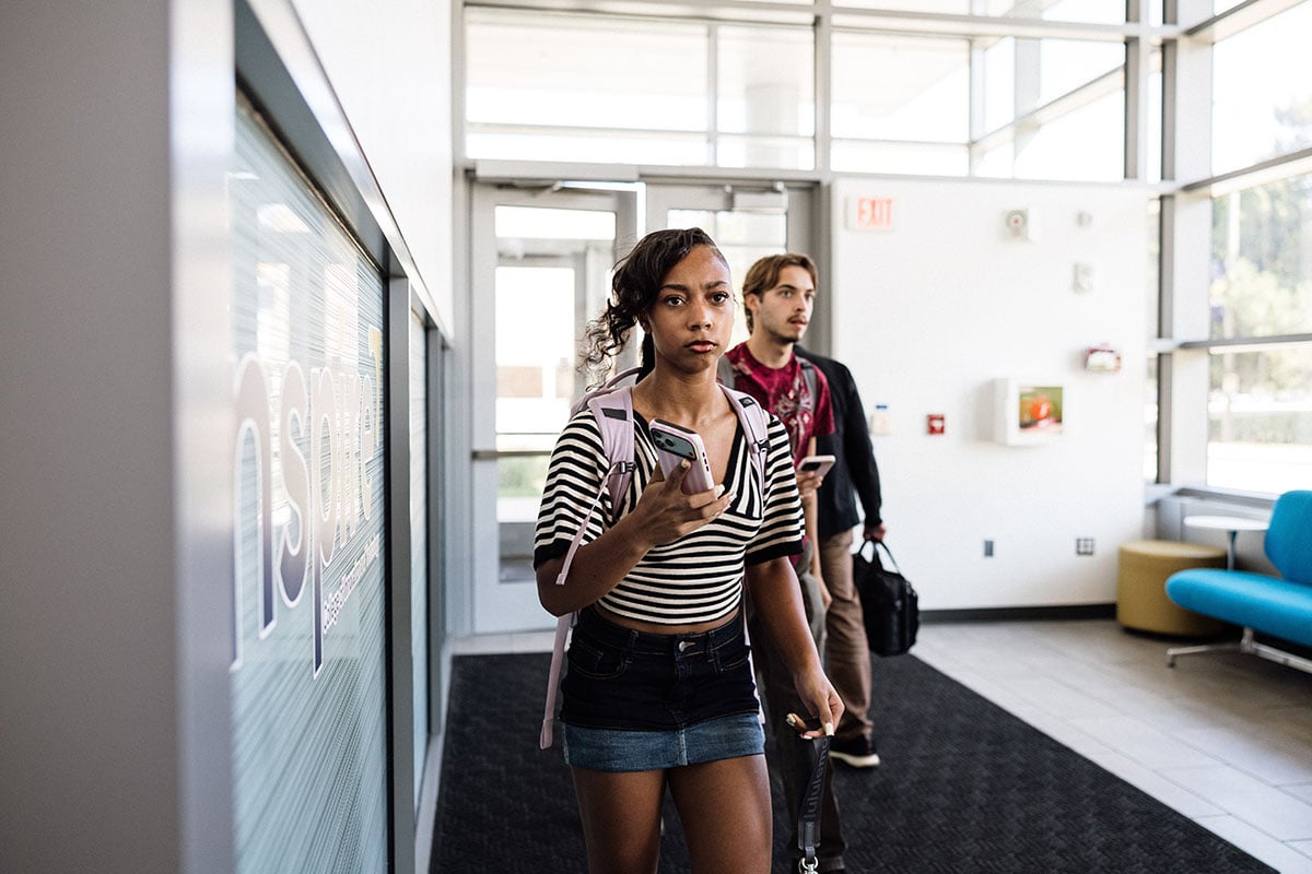 FLINT_First Day of Class Fall 2026_0008 A young girl with curly hair, wearing a striped shirt and denim shorts, walks through a modern building holding a smartphone. A young man follows behind her, also looking at his phone.