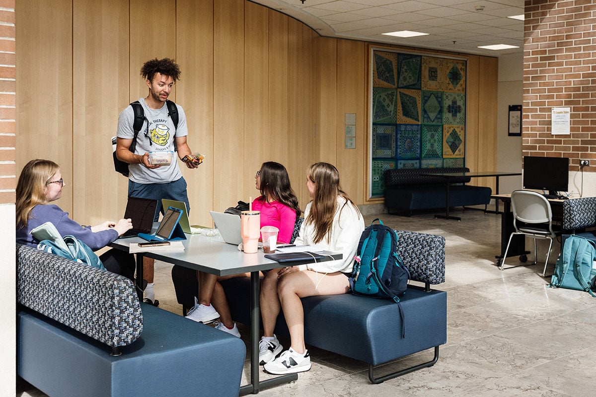 FLINT_First Day of Class Fall 2026_0017 A group of four students in a study area, with one male student standing and talking while holding snacks, and three female students seated at a table with laptops and drinks.