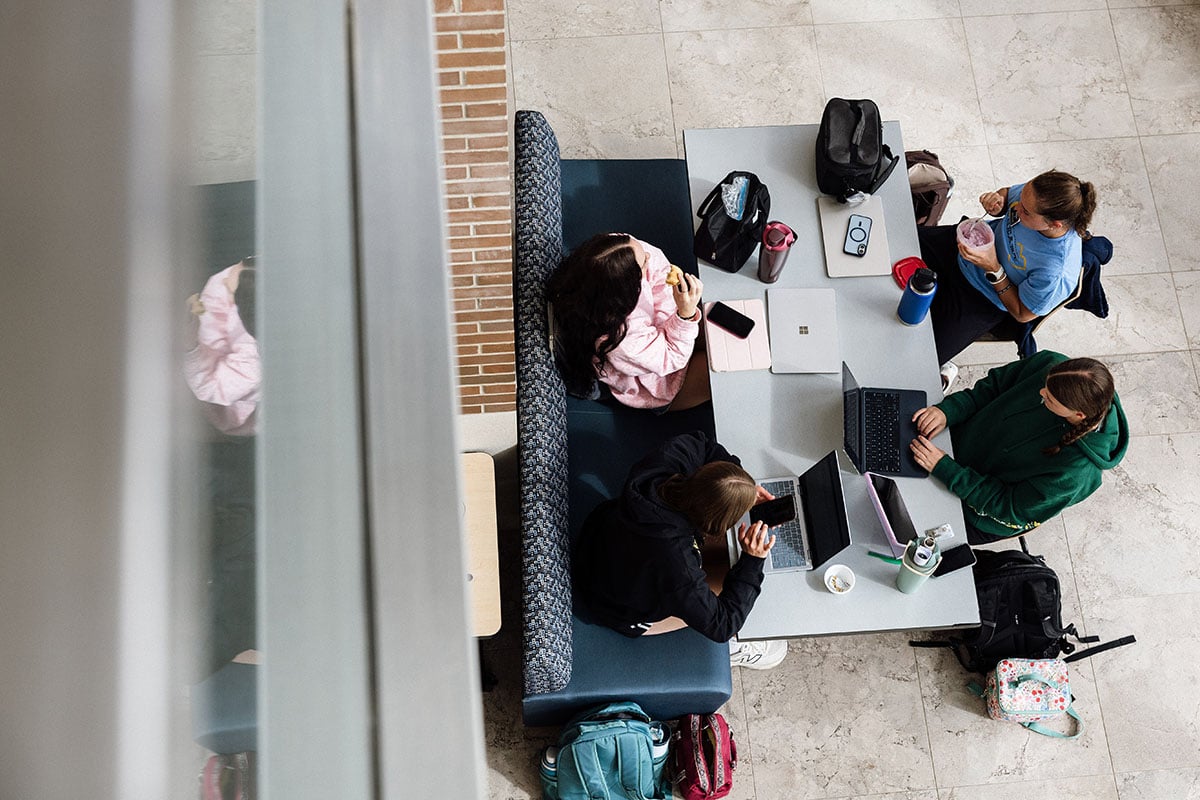 FLINT_First Day of Class Fall 2026_0019 Aerial view of four students sitting around a table, working on laptops and using phones, with backpacks and drinks nearby.