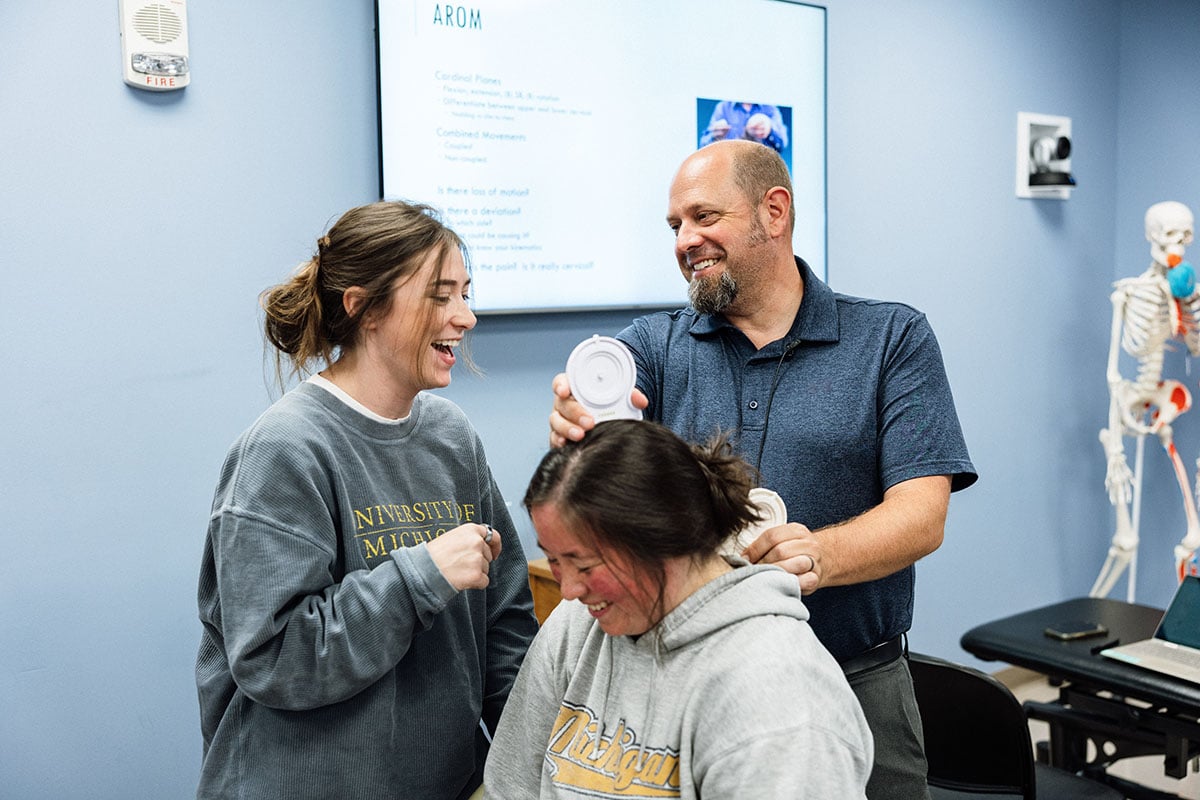 FLINT_First Day of Class Fall 2026_0026 A man is smiling and demonstrating a technique on a woman sitting in front of him, while another woman laughs beside them. A presentation screen is visible in the background.