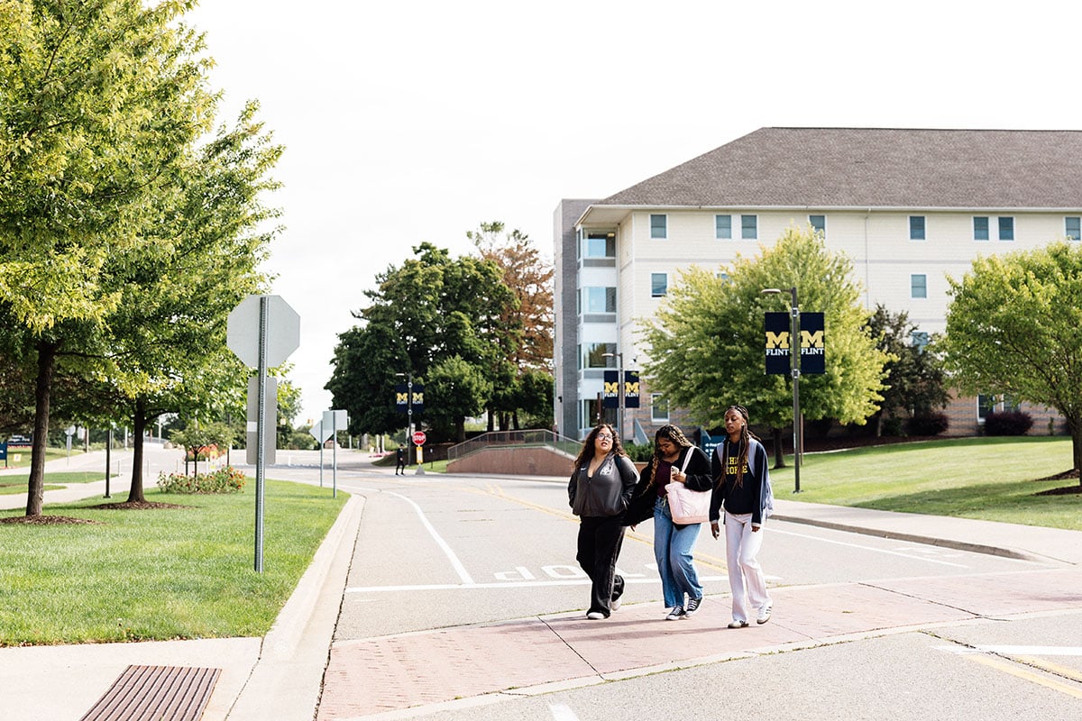 FLINT_First Day of Class Fall 2026_0027 Three students walking together across a crosswalk in front of a university building, with trees and signage in the background.