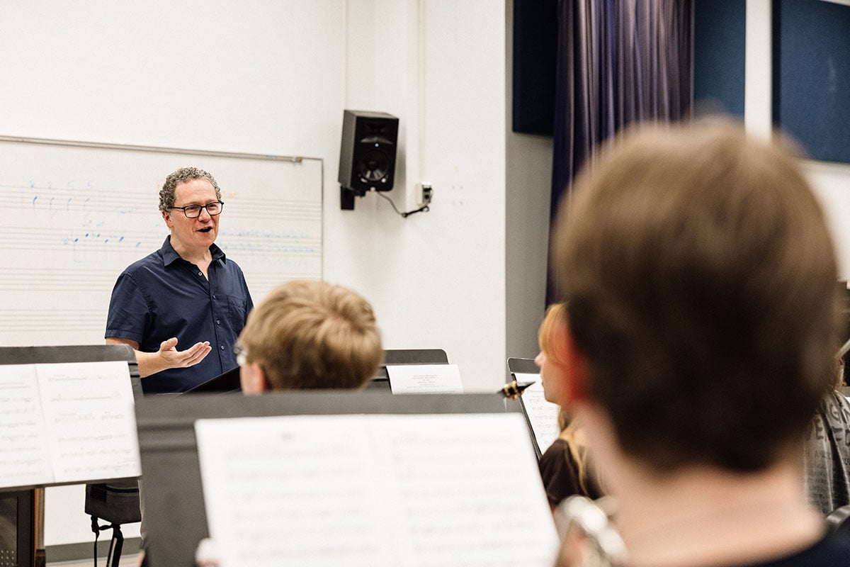 FLINT_First Day of Class Fall 2026_0028 A music instructor stands in front of a group of students, gesturing while speaking during a rehearsal. Music sheets are visible in the foreground.