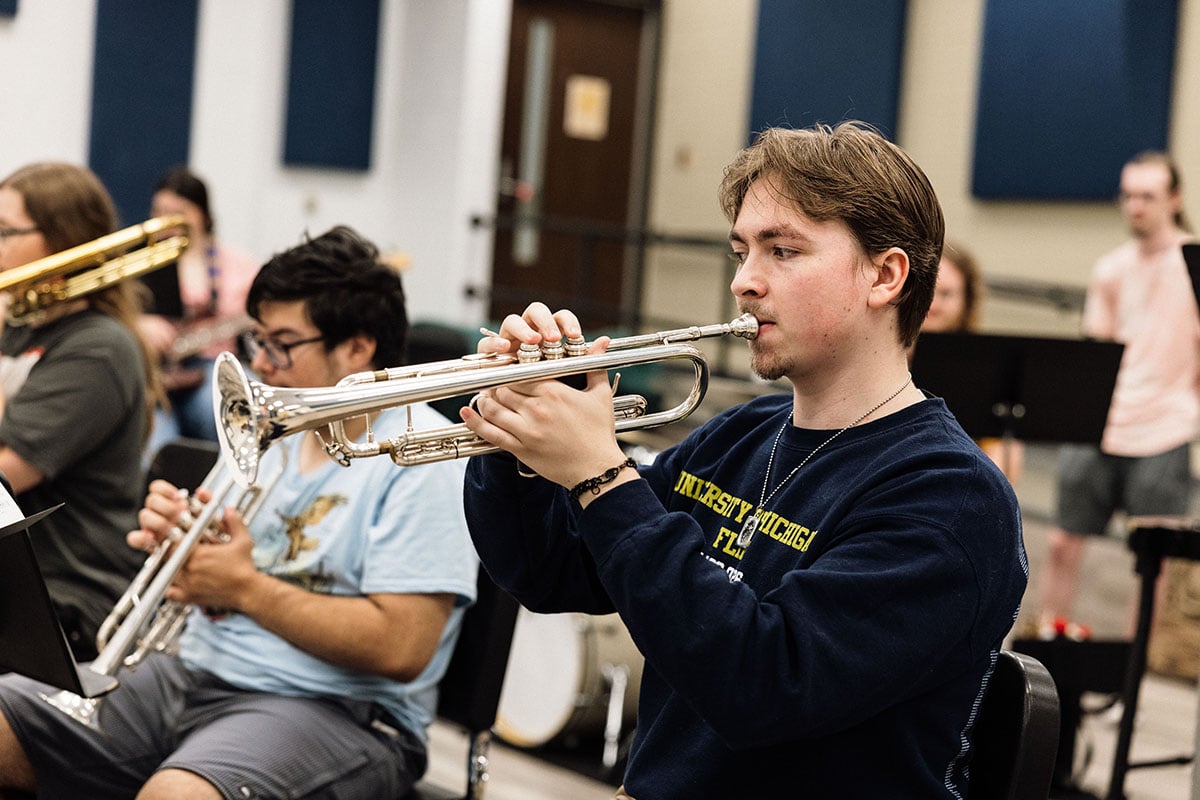 FLINT_First Day of Class Fall 2026_0030 A young man plays a trumpet in a music rehearsal setting, surrounded by other musicians. He wears a dark sweatshirt and is focused on his instrument.
