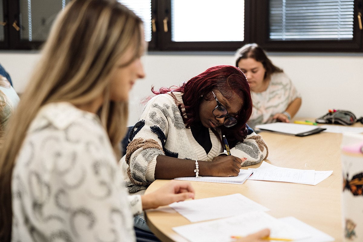 FLINT_First Day of Class Fall 2026_0034 A group of students in a classroom setting, with one student focused on writing notes at a table, while others are engaged in their work.